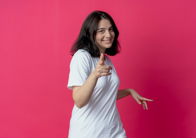 Smiling young pretty caucasian girl pointing at camera and keeping hand in air isolated on crimson background with copy space