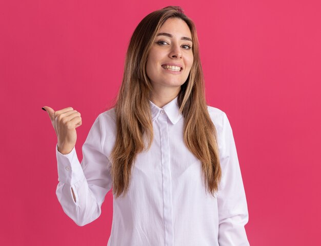 Smiling young pretty caucasian girl points at side on pink 