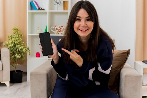 Smiling young pretty caucasian girl sitting on armchair in designed living room showing mobile phone  pointing at it and 