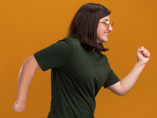 Smiling young pretty caucasian girl in sun glasses stands sideways pretending to run on orange