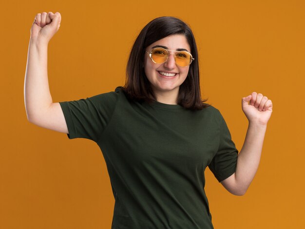 Smiling young pretty caucasian girl in sun glasses stands with raised fists on orange