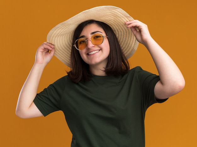 Улыбающаяся молодая симпатичная кавказская девушка в солнцезащитных очках и в пляжной шляпе, изолированной на оранжевой стене с копией пространства