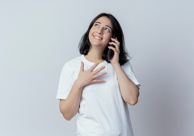 Улыбающаяся молодая симпатичная кавказская девушка разговаривает по телефону и смотрит вверх изолированно на белом фоне с копией пространства