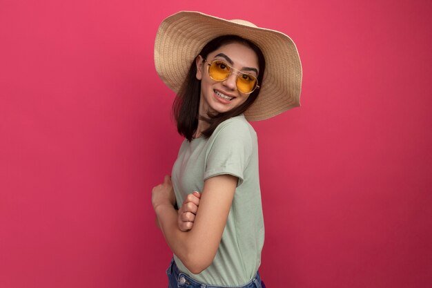 Smiling young pretty caucasian girl wearing beach hat and sunglasses standing with closed posture in profile view isolated on pink wall with copy space