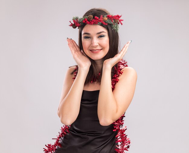 Smiling young pretty caucasian girl wearing christmas head wreath and tinsel garland around neck looking at camera keeping hands near head isolated on white background with copy space