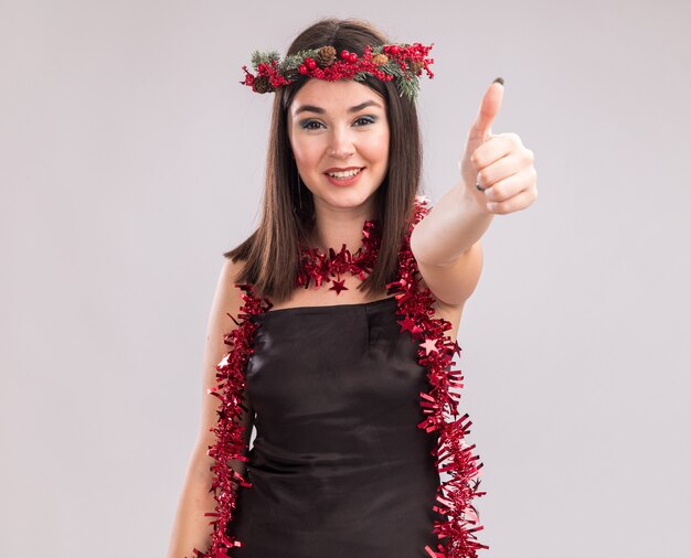 クリスマスの頭の花輪と首の周りに見掛け倒しの花輪を身に着けている若いかわいい白人の女の子の笑顔は、コピースペースで白い背景に分離された親指を示すカメラを見て