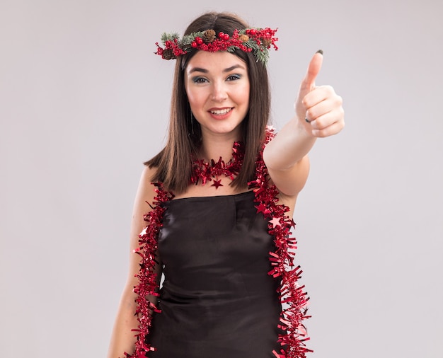 クリスマスの頭の花輪と首の周りに見掛け倒しの花輪を身に着けている若いかわいい白人の女の子の笑顔は、コピースペースで白い背景に分離された親指を示すカメラを見て