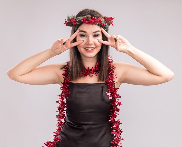 Smiling young pretty caucasian girl wearing christmas head wreath and tinsel garland around neck looking at camera showing v-sign symbols near eyes isolated on white background