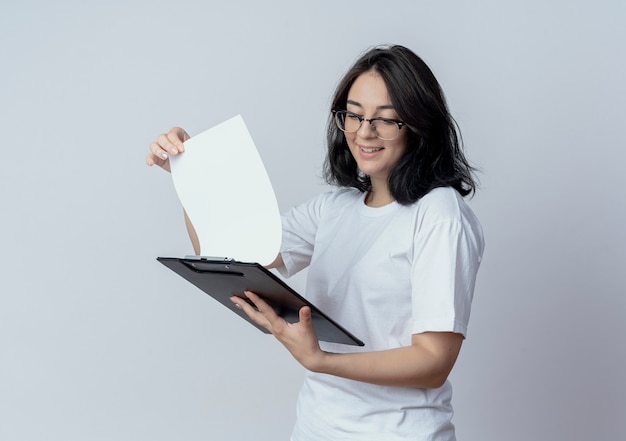 Smiling young pretty caucasian girl wearing glasses holding and looking at clipboard isolated on white background