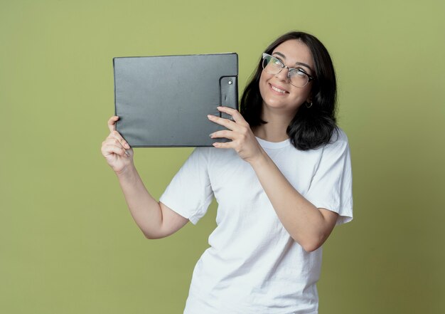 Smiling young pretty caucasian girl wearing glasses looking up holding clipboard  on olive green space