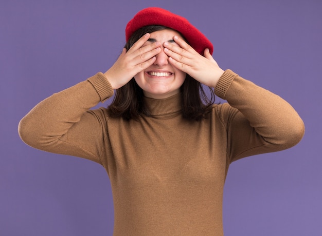 Smiling young pretty caucasian girl with beret hat covers eyes with hands isolated on purple wall with copy space