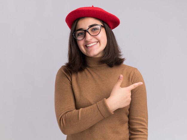 Smiling young pretty caucasian girl with beret hat and in optical glasses pointing at side isolated on white wall with copy space