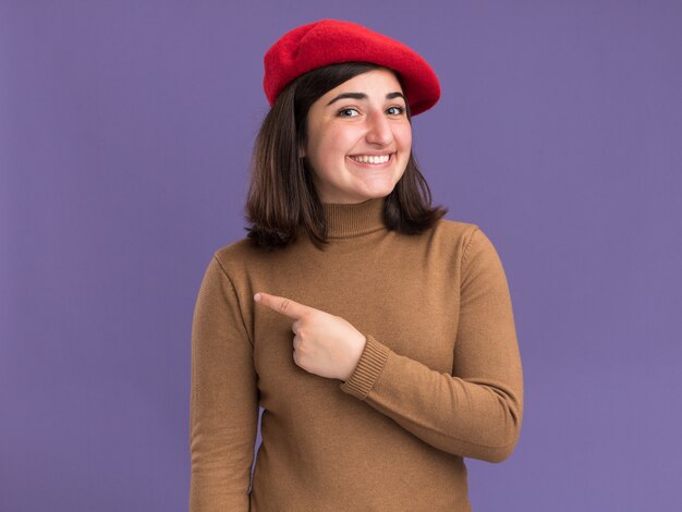 コピースペースと紫色の壁に分離された側を指しているベレー帽の帽子と笑顔の若いかなり白人の女の子