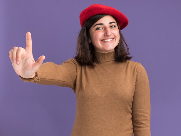 コピースペースと紫色の壁に分離された上向きのベレー帽の帽子と笑顔の若いかなり白人の女の子