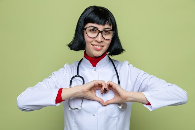 Smiling young pretty caucasian girl with optical glasses in doctor uniform with stethoscope gesturing heart sign