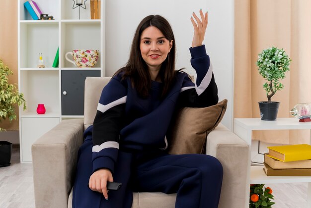 Smiling young pretty caucasian woman sitting on armchair in designed living room holding remote control looking keeping hand in air