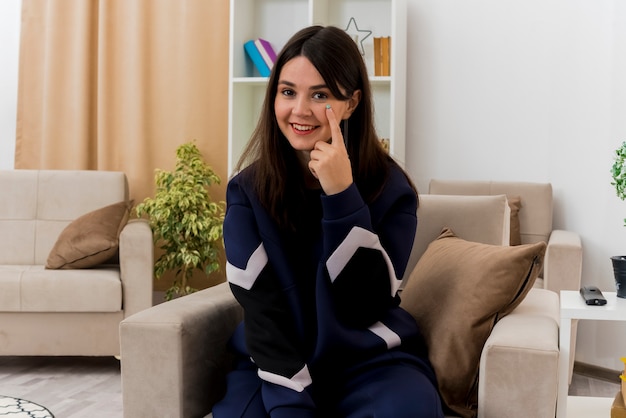 Smiling young pretty caucasian woman sitting on armchair in designed living room looking putting finger under eye