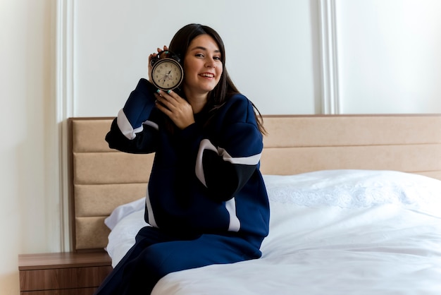 Smiling young pretty caucasian woman sitting on bed in bedroom holding alarm clock near head looking