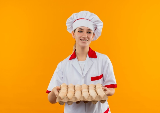 Sorridente giovane cuoco grazioso in uniforme da chef con parentesi graffe dentali che tiene il cartone di uova isolato su spazio arancione