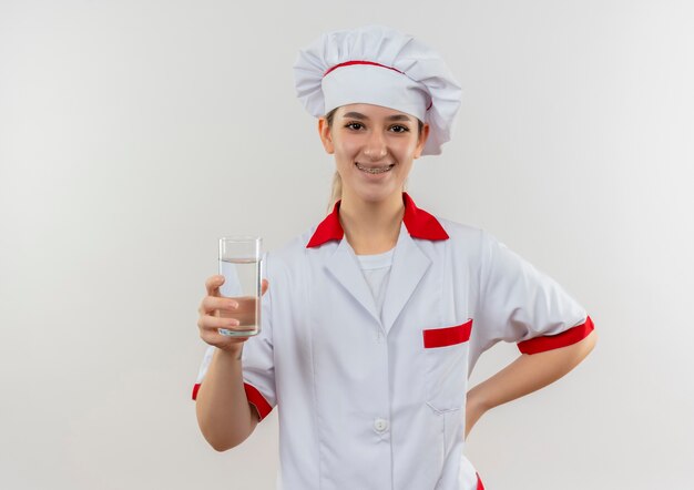 물 한 잔을 들고 흰색 공간에 고립 된 허리에 손을 넣어 치과 교정기와 요리사 유니폼에 젊은 예쁜 요리사 미소