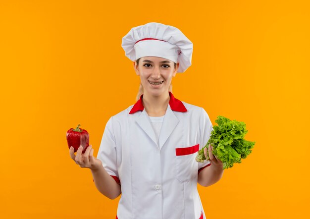 Smiling young pretty cook in chef uniform with dental braces holding pepper and lettuce isolated on orange space