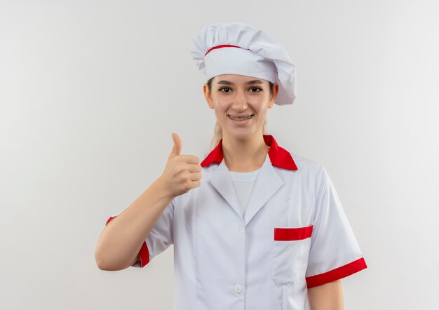 笑顔の若いかわいい料理人がシェフの制服を着て、白いスペースで孤立しているように見える親指を示す歯列矯正器