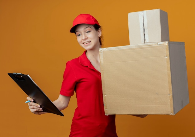 Бесплатное фото Улыбающаяся молодая симпатичная доставщица в красной форме и кепке, держащая ручку и буфер обмена, смотрит в буфер обмена с картонными коробками в другой руке, изолированной на оранжевом фоне