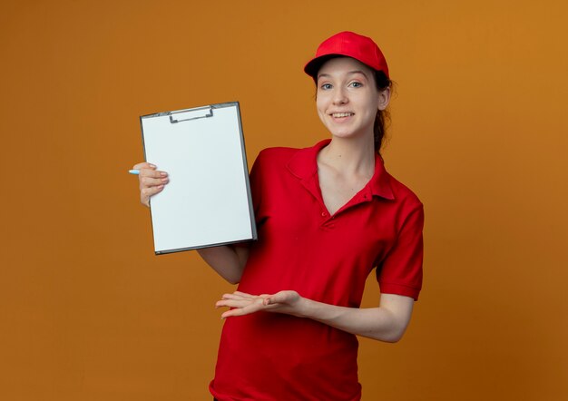 Smiling young pretty delivery girl in red uniform and cap holding pen and showing clipboard and pointing with hand at clipboard isolated on orange background with copy space