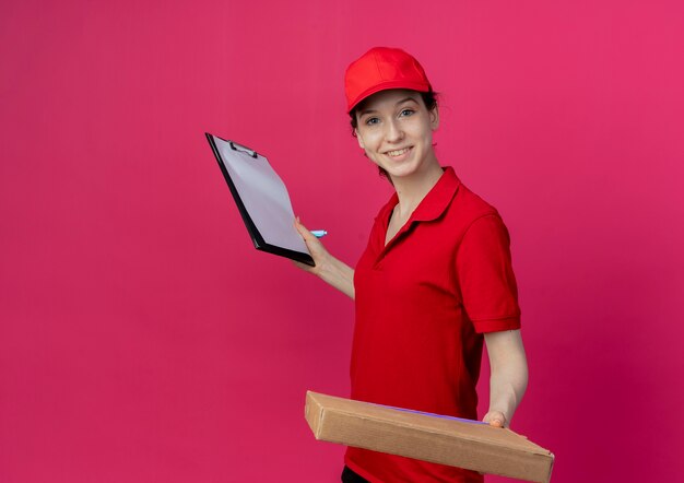 빨간 유니폼과 모자 복사 공간이 진홍색 배경에 고립 된 펜으로 피자 패키지와 클립 보드를 들고 웃는 젊은 예쁜 배달 소녀