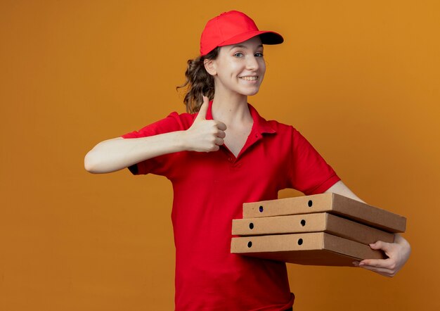 Улыбающаяся молодая симпатичная доставщица в красной форме и кепке держит пакеты с пиццей и показывает большой палец вверх, изолированные на оранжевом фоне