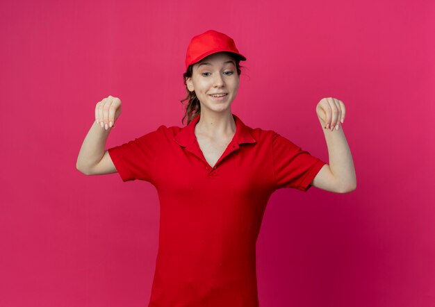 빨간 제복을 입은 젊은 예쁜 배달 소녀 미소 짓고 모자를 내려다보고 진홍색 배경에 고립 된 뭔가를 들고 척
