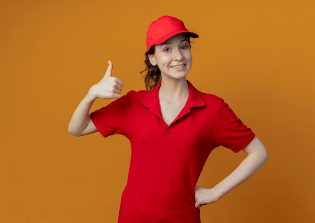 Smiling young pretty delivery girl in red uniform and cap putting hand on waist and showing thumb up isolated on orange background with copy space