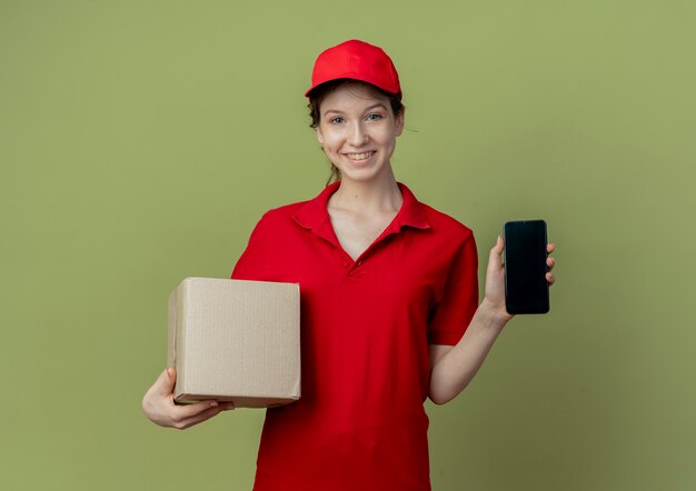 Smiling young pretty delivery girl in red uniform and cap showing mobile phone and holding carton box isolated on olive green background