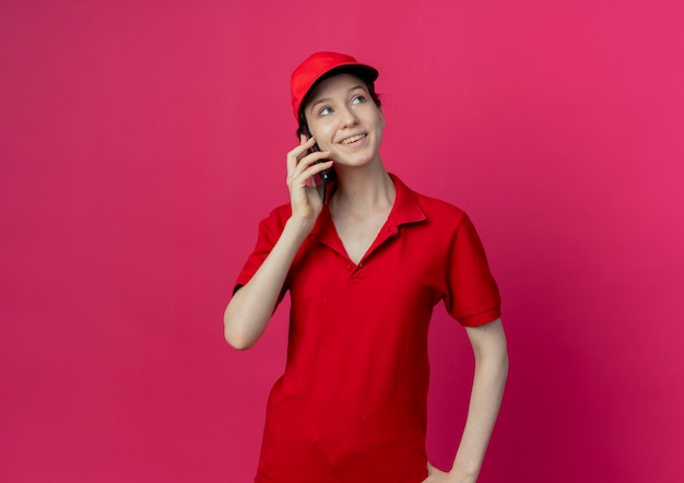빨간 유니폼과 모자 복사 공간이 진홍색 배경에 고립 된 측면을보고 전화로 얘기하는 젊은 예쁜 배달 소녀 미소