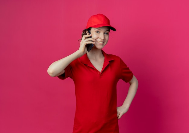 Smiling young pretty delivery girl in red uniform and cap talking on phone putting hand on waist isolated on crimson background with copy space