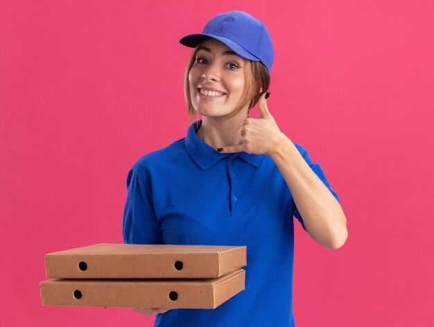 Улыбающаяся молодая симпатичная доставщица в униформе висят свободно и держит коробки для пиццы на розовом