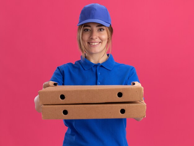 Smiling young pretty delivery girl in uniform holding pizza boxes on pink