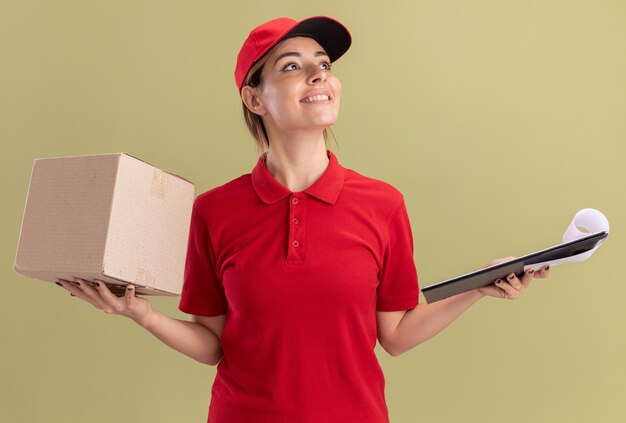 Улыбающаяся молодая симпатичная доставщица в униформе держит буфер обмена и картонную коробку, глядя в сторону на оливково-зеленом