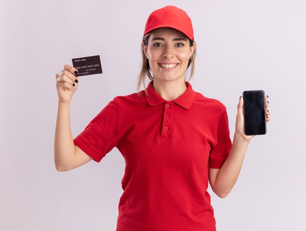 Улыбающаяся молодая симпатичная доставщица в униформе держит кредитную карту и телефон на белом