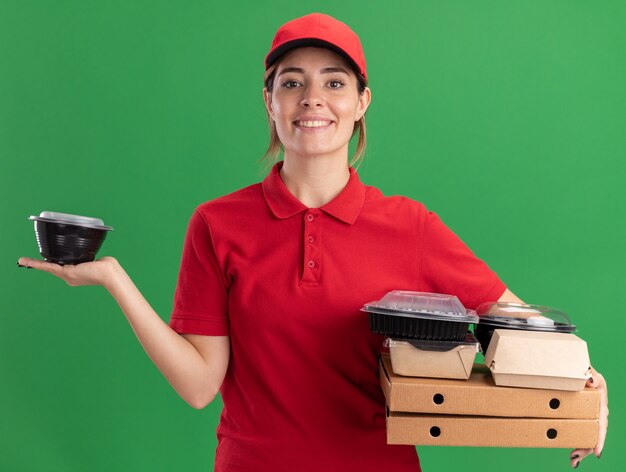 Smiling young pretty delivery girl in uniform holds paper food packages and containers on pizza boxes looking at camera on green