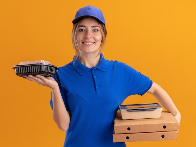 Улыбающаяся молодая симпатичная доставщица в униформе держит бумажные пакеты с едой и контейнеры на коробках для пиццы на оранжевом