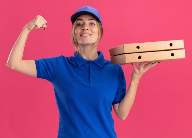 Smiling young pretty delivery girl in uniform holds pizza boxes and tenses biceps on pink