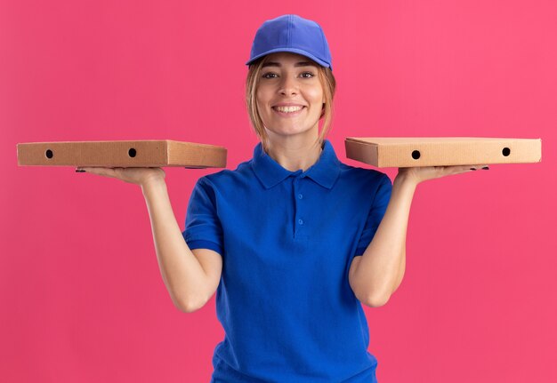 Smiling young pretty delivery girl in uniform holds pizza boxes on two hands on pink