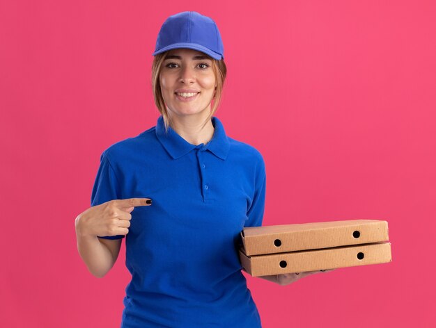 Smiling young pretty delivery girl in uniform holds and points at pizza boxes on pink