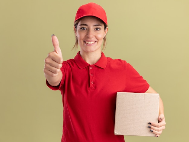 Улыбающаяся молодая симпатичная доставщица в униформе показывает палец вверх и держит картонную коробку на оливково-зеленом