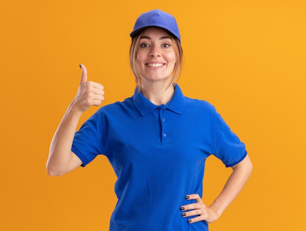 Smiling young pretty delivery girl in uniform thumbs up looking at camera on orange