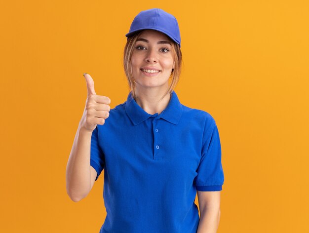 Smiling young pretty delivery girl in uniform thumbs up on orange