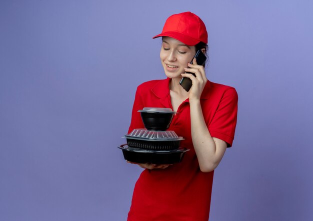 Smiling young pretty delivery girl wearing red uniform and cap holding and looking at food containers and talking on phone