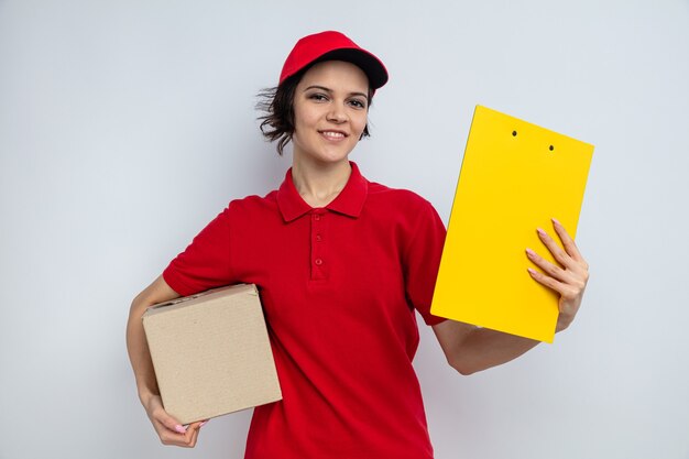 Smiling young pretty delivery woman holding cardboard box and clipboard 