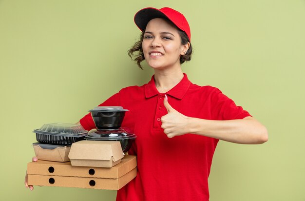 Smiling young pretty delivery woman holding food containers with packaging on pizza boxes and thumbing up 